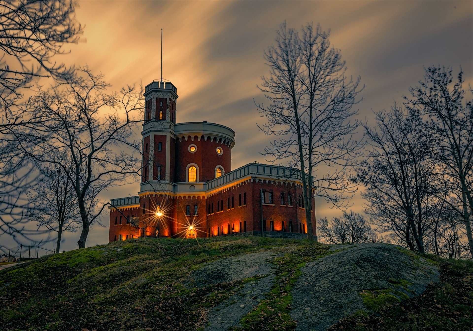 Kastellholmen Castle in Sweden illuminated at dusk, set against a dramatic sky with bare trees surrounding the historic man-made structure.