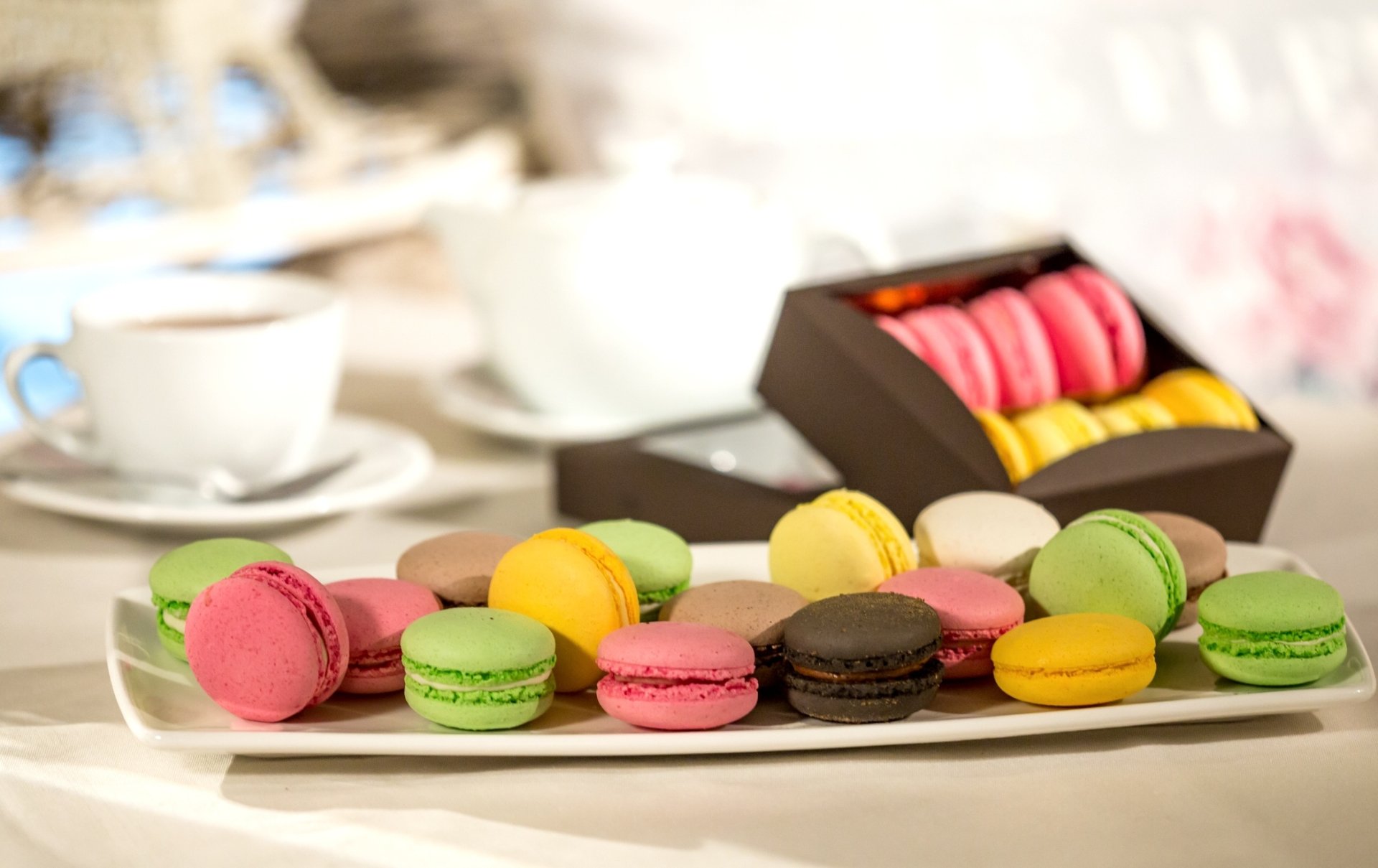 HD desktop wallpaper featuring a still life of colorful macarons arranged on a plate, with a box of sweets and blurred teacups in the background.