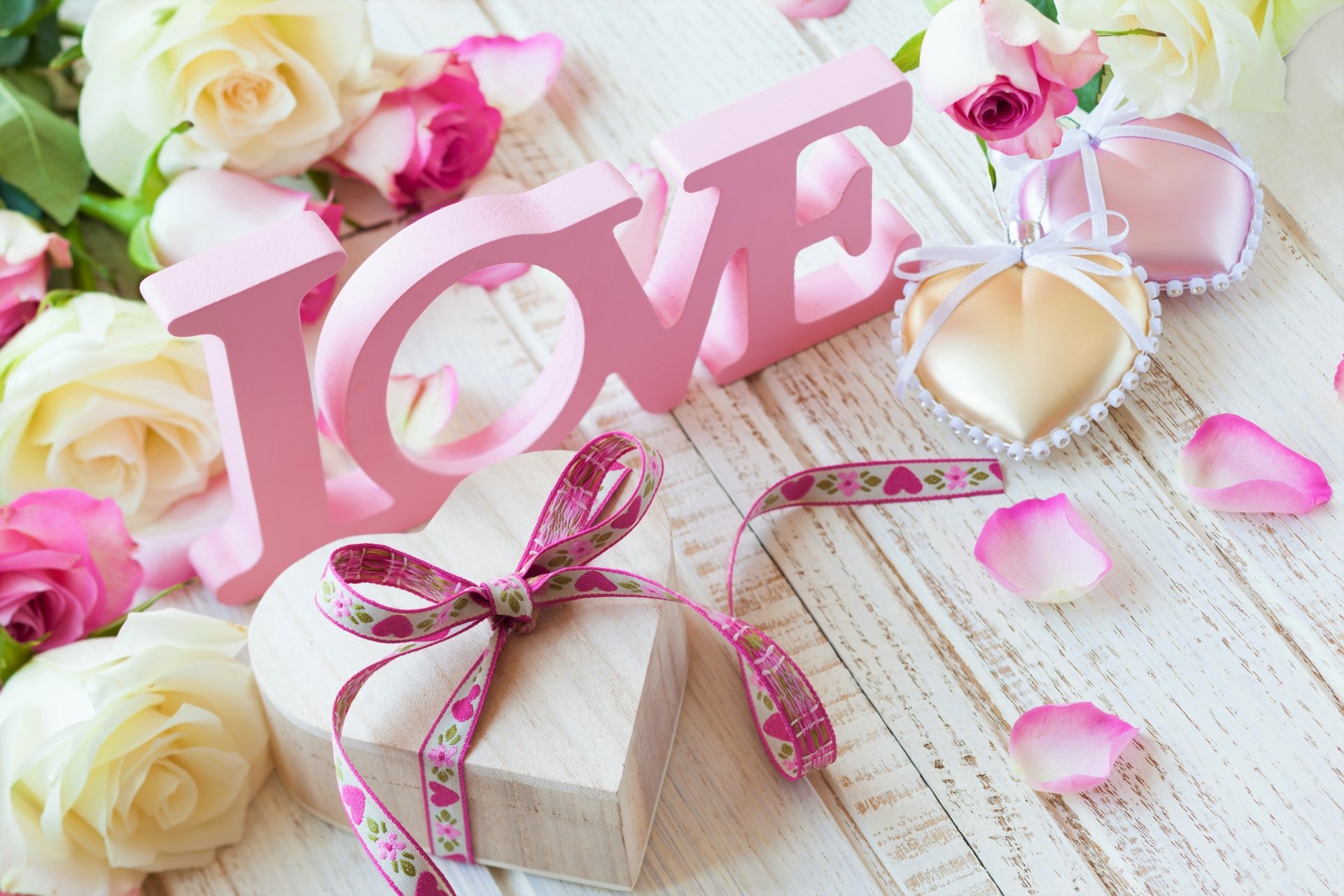 HD wallpaper featuring a pink LOVE sign, a heart-shaped box with a ribbon, roses, and scattered petals on a wooden background, creating a romantic and artistic atmosphere.
