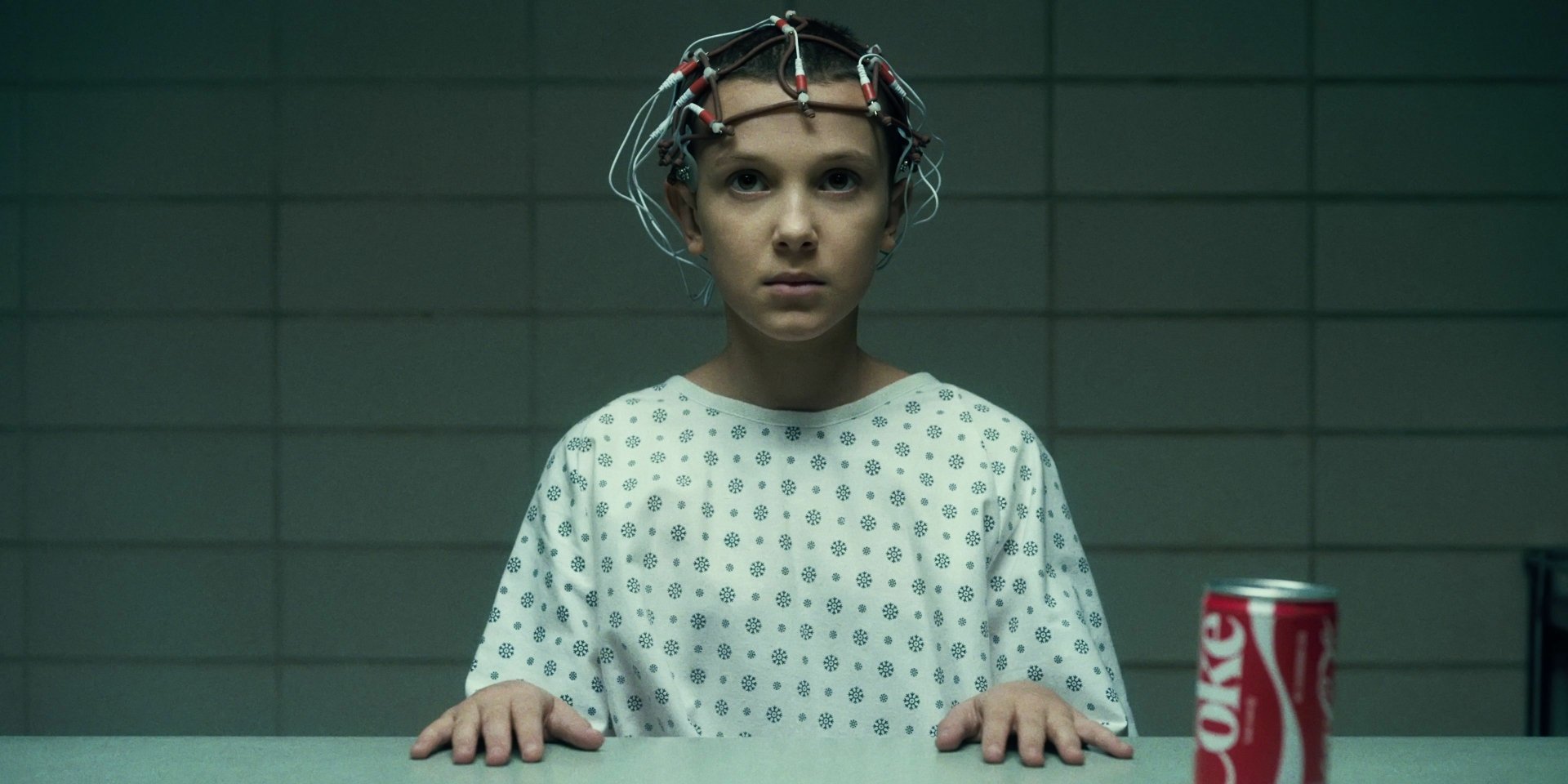 Eleven from Stranger Things, played by Millie Bobby Brown, wearing a hospital gown and a neural monitoring device, with a can of Diet Coke on the table in this HD PC desktop wallpaper.