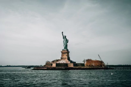 HD desktop wallpaper of the man-made Statue of Liberty in the USA, standing tall against a cloudy sky with water in the foreground.