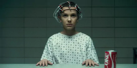 Eleven from Stranger Things, played by Millie Bobby Brown, wearing a hospital gown and a neural monitoring device, with a can of Diet Coke on the table in this HD PC desktop wallpaper.