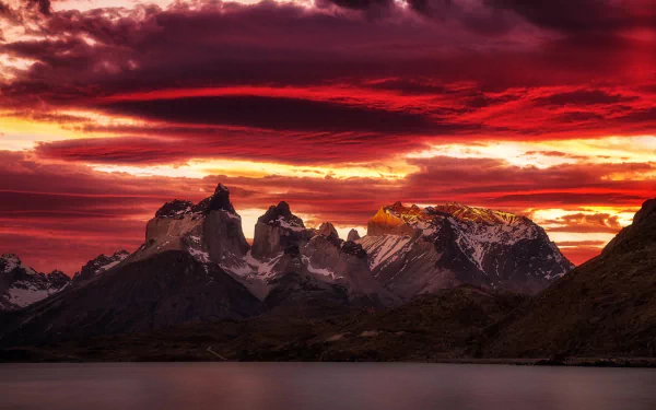 HD desktop wallpaper of a mountain peak at sunset with a dramatic sky filled with vibrant red and orange clouds, reflecting on a tranquil body of water below.