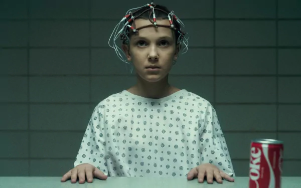 Eleven from Stranger Things, played by Millie Bobby Brown, wearing a hospital gown and a neural monitoring device, with a can of Diet Coke on the table in this HD PC desktop wallpaper.