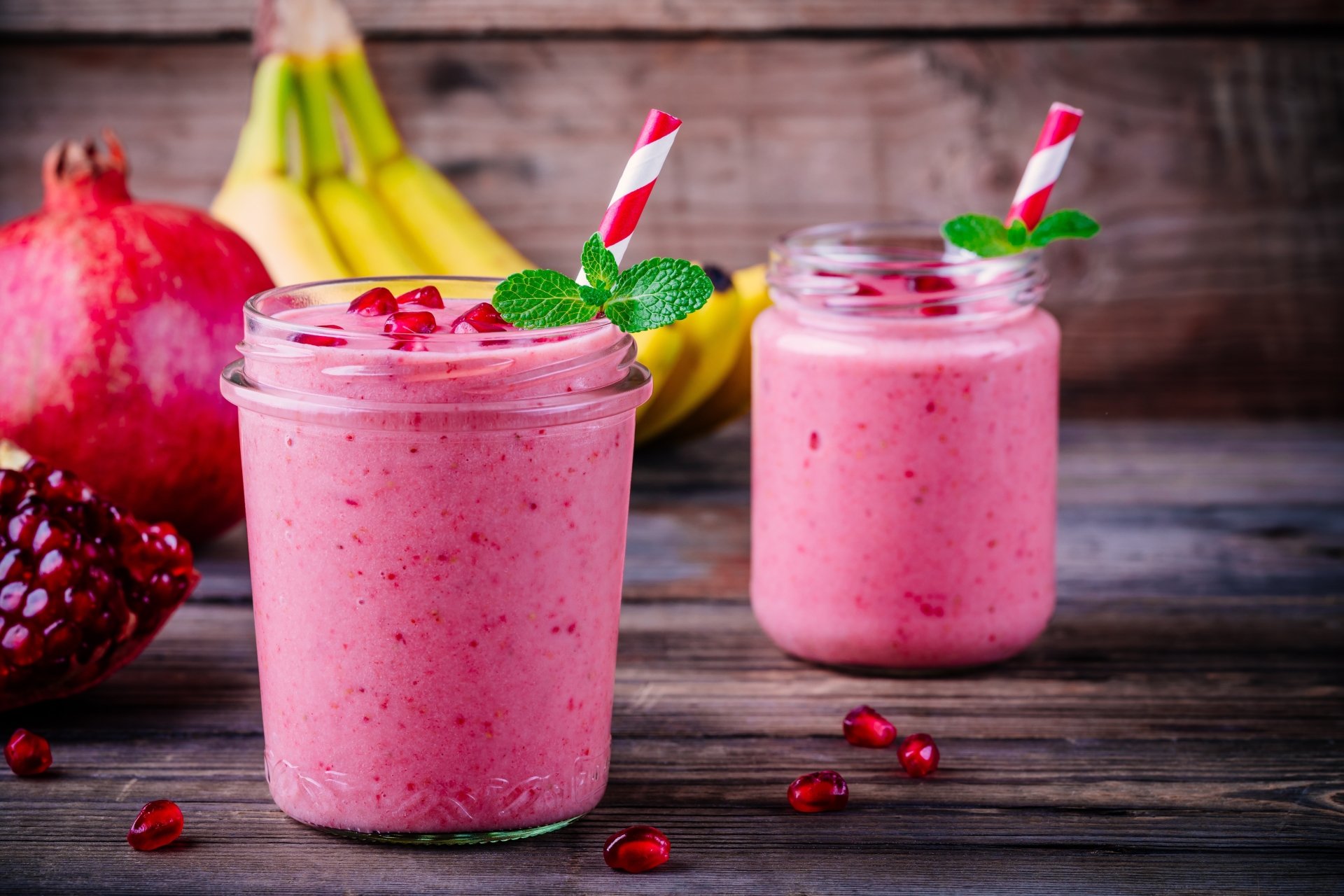 8K Ultra HD wallpaper showing two jars of pink pomegranate smoothie with mint and striped straws, fresh pomegranate and bananas on a rustic wooden surface.