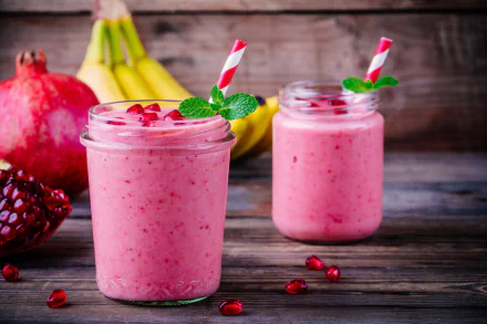 8K Ultra HD wallpaper showing two jars of pink pomegranate smoothie with mint and striped straws, fresh pomegranate and bananas on a rustic wooden surface.