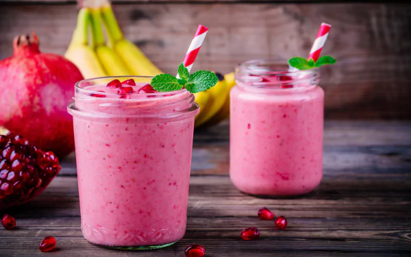 8K Ultra HD wallpaper showing two jars of pink pomegranate smoothie with mint and striped straws, fresh pomegranate and bananas on a rustic wooden surface.
