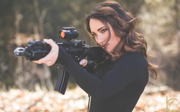 Brunette model Cheyenne Mykel aims a Colt AR-15 assault rifle with orange eyes in an outdoor setting, featured in a Girls & Guns HD desktop wallpaper.