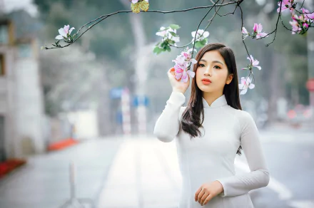 HD PC wallpaper of an Asian woman model with long black hair and brown eyes, standing near blossoming branches with a soft depth of field background.