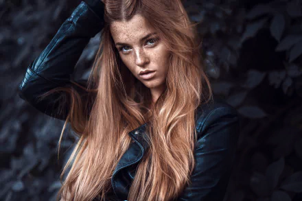 Close-up HD desktop wallpaper of a freckled redhead model with long flowing hair and a leather jacket against moody foliage background.