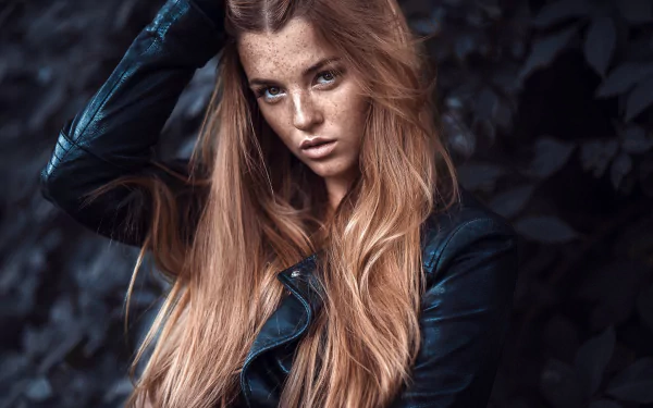 Close-up HD desktop wallpaper of a freckled redhead model with long flowing hair and a leather jacket against moody foliage background.