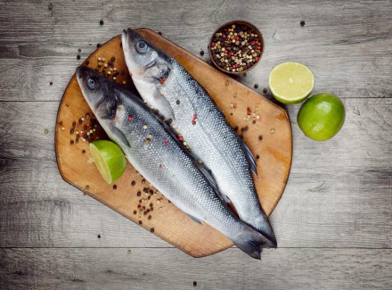 A 4K Ultra HD still life of two whole fish on a wooden board, garnished with lime wedges and scattered mixed peppercorns, set against a rustic wooden background.