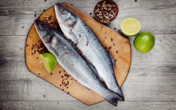 A 4K Ultra HD still life of two whole fish on a wooden board, garnished with lime wedges and scattered mixed peppercorns, set against a rustic wooden background.