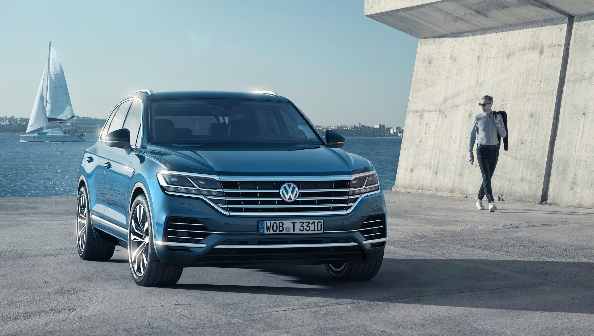 Blue Volkswagen Touareg SUV parked on a waterfront concrete promenade with sailboats and a pedestrian in view — 4K Ultra HD PC desktop wallpaper.