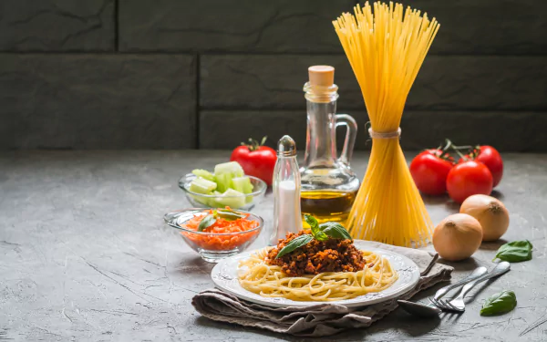 HD desktop wallpaper featuring a still life of a pasta meal with spaghetti, sauce, fresh tomatoes, onions, olive oil, and seasonings arranged on a gray textured surface.