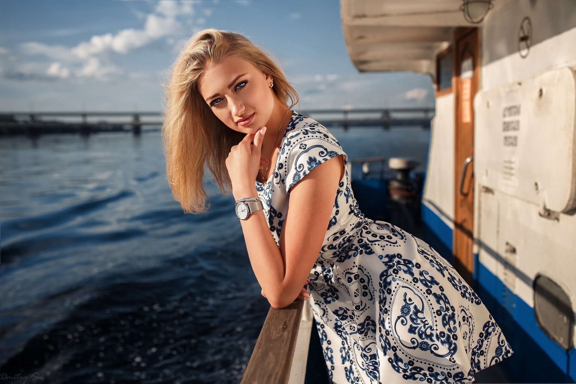 Blonde woman with blue eyes and a patterned dress leans on a boat railing, captured with depth of field in an HD desktop wallpaper background.
