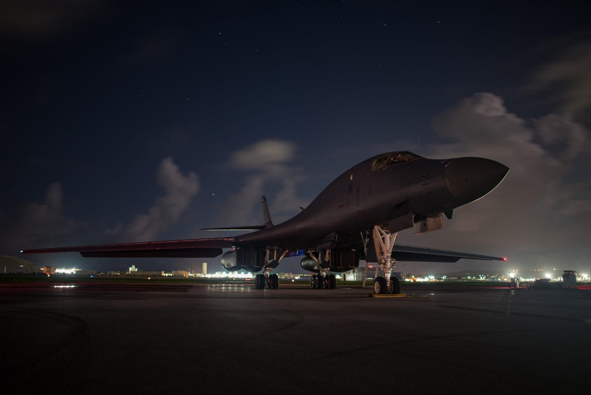 HD wallpaper of a Rockwell B-1 Lancer warplane on the tarmac at night, showcasing the military bomber's sleek design and powerful presence against a backdrop of distant city lights and a night sky.
