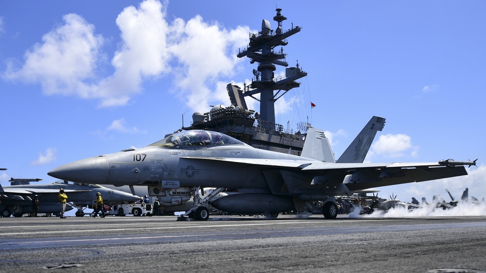 HD desktop wallpaper featuring a Boeing F/A-18E/F Super Hornet jet fighter warplane on the deck of an aircraft carrier under a clear blue sky.