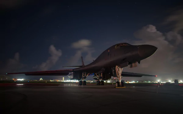 HD wallpaper of a Rockwell B-1 Lancer warplane on the tarmac at night, showcasing the military bomber's sleek design and powerful presence against a backdrop of distant city lights and a night sky.