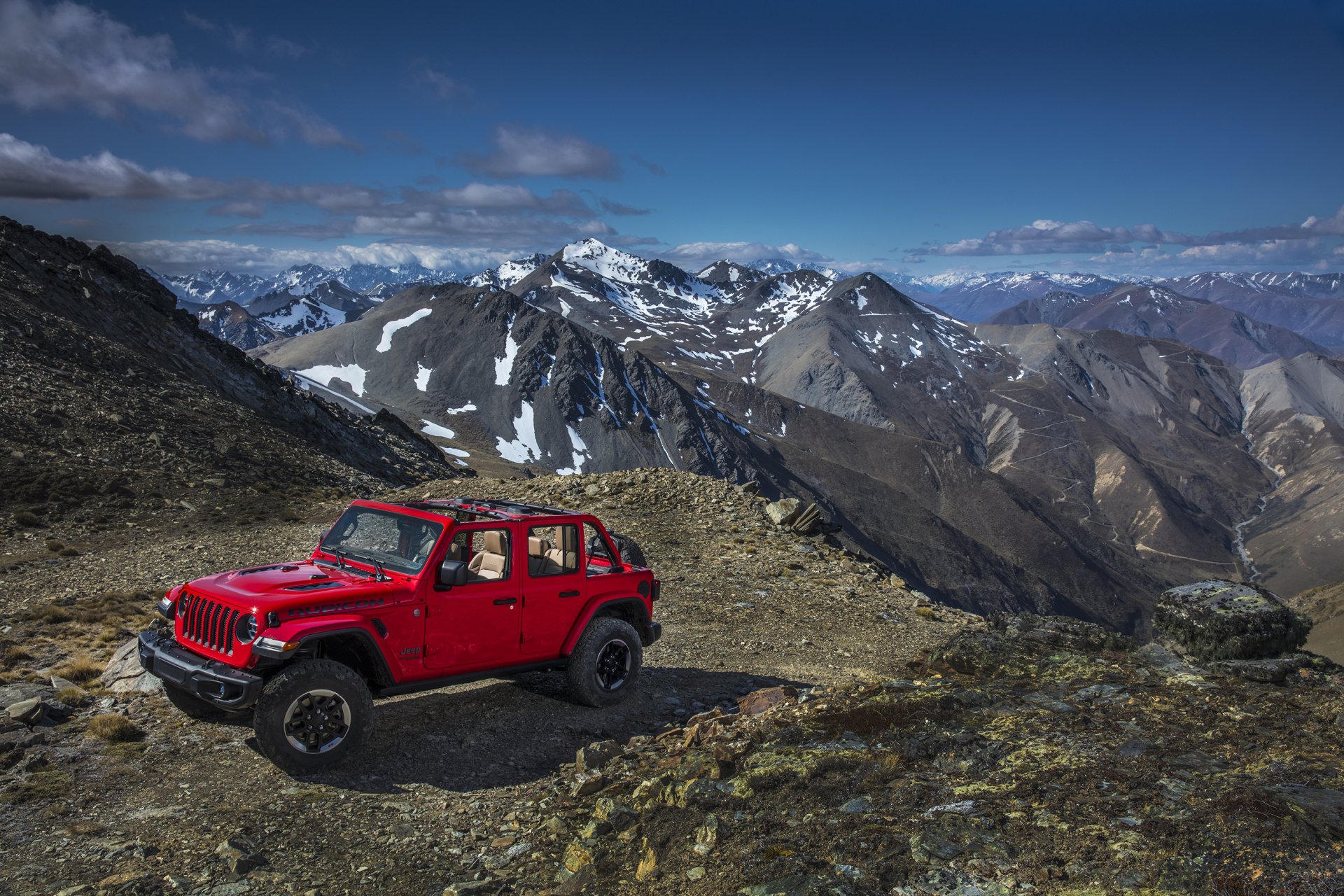 Red Jeep Wrangler off-road vehicle parked on rugged mountain terrain with snowy peaks in the background, captured in an HD PC desktop wallpaper.