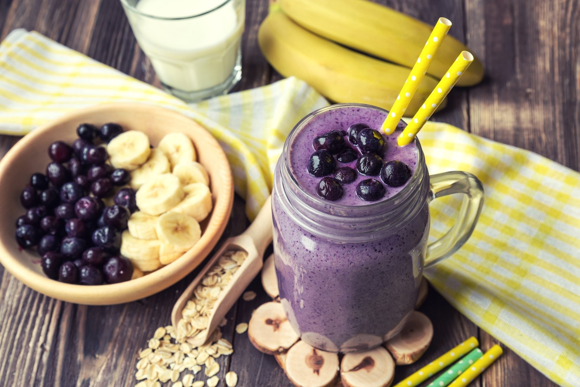 HD desktop wallpaper featuring a blueberry and banana smoothie in a glass mug, surrounded by fresh blueberries, banana slices, oats, and a glass of milk in a still life arrangement.