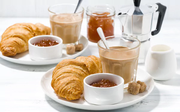 A 4K Ultra HD desktop wallpaper featuring a breakfast scene with golden croissants, a cup of coffee with milk, jars of jam, and a moka pot on a white table.