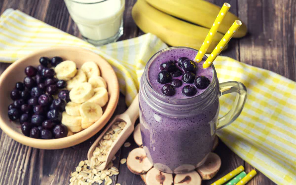 HD desktop wallpaper featuring a blueberry and banana smoothie in a glass mug, surrounded by fresh blueberries, banana slices, oats, and a glass of milk in a still life arrangement.
