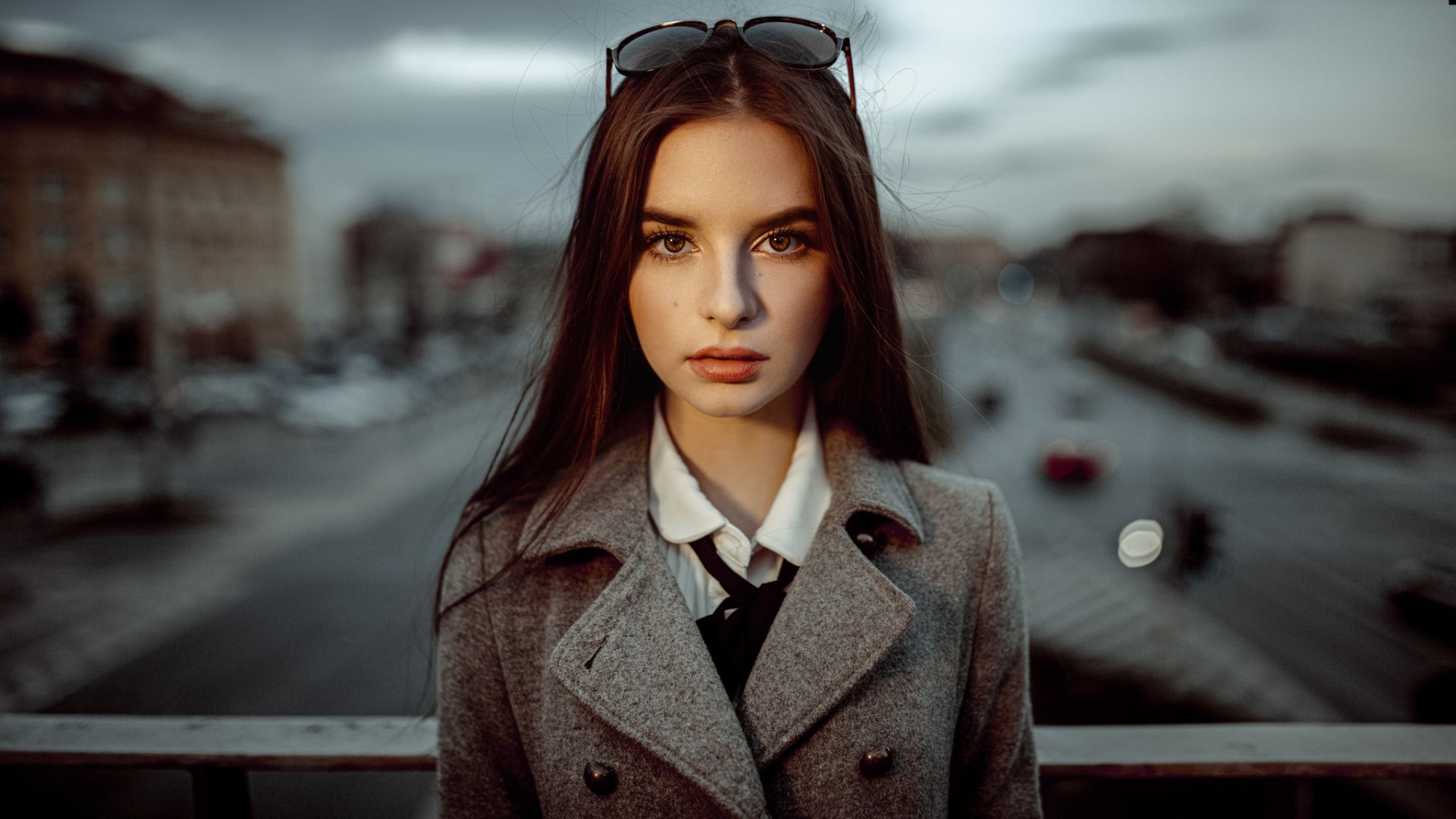 HD PC desktop wallpaper: close-up of a brunette model’s face with brown eyes and sunglasses on her head, shallow depth-of-field and blurred urban background.