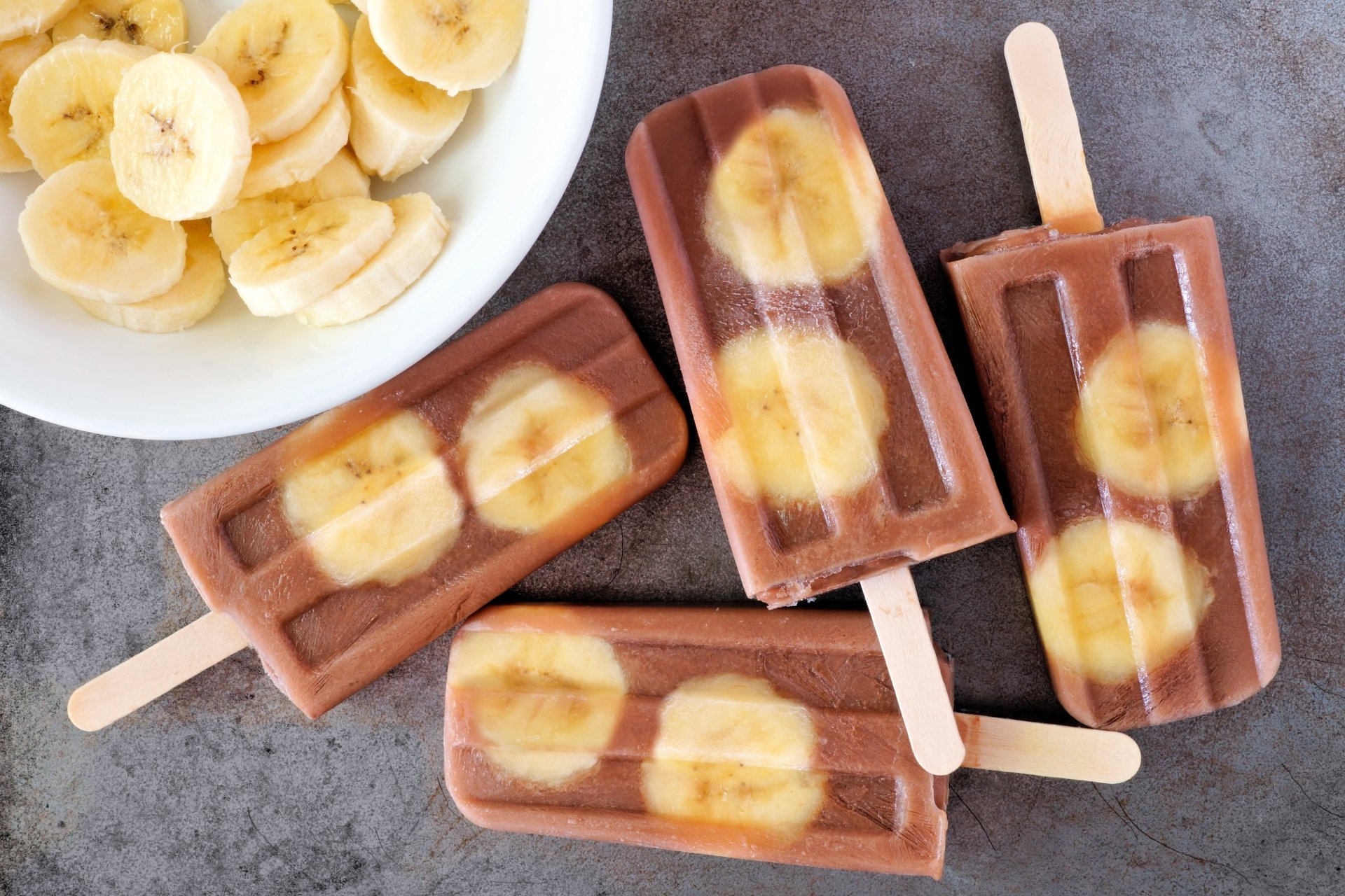 A 4K Ultra HD desktop wallpaper showing chocolate banana ice cream popsicles with banana slices on a gray surface alongside a bowl of sliced bananas.