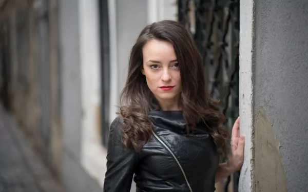 8K Ultra HD wallpaper of a brunette woman model with brown eyes and lipstick, wearing a leather jacket against a shallow depth-of-field urban background.