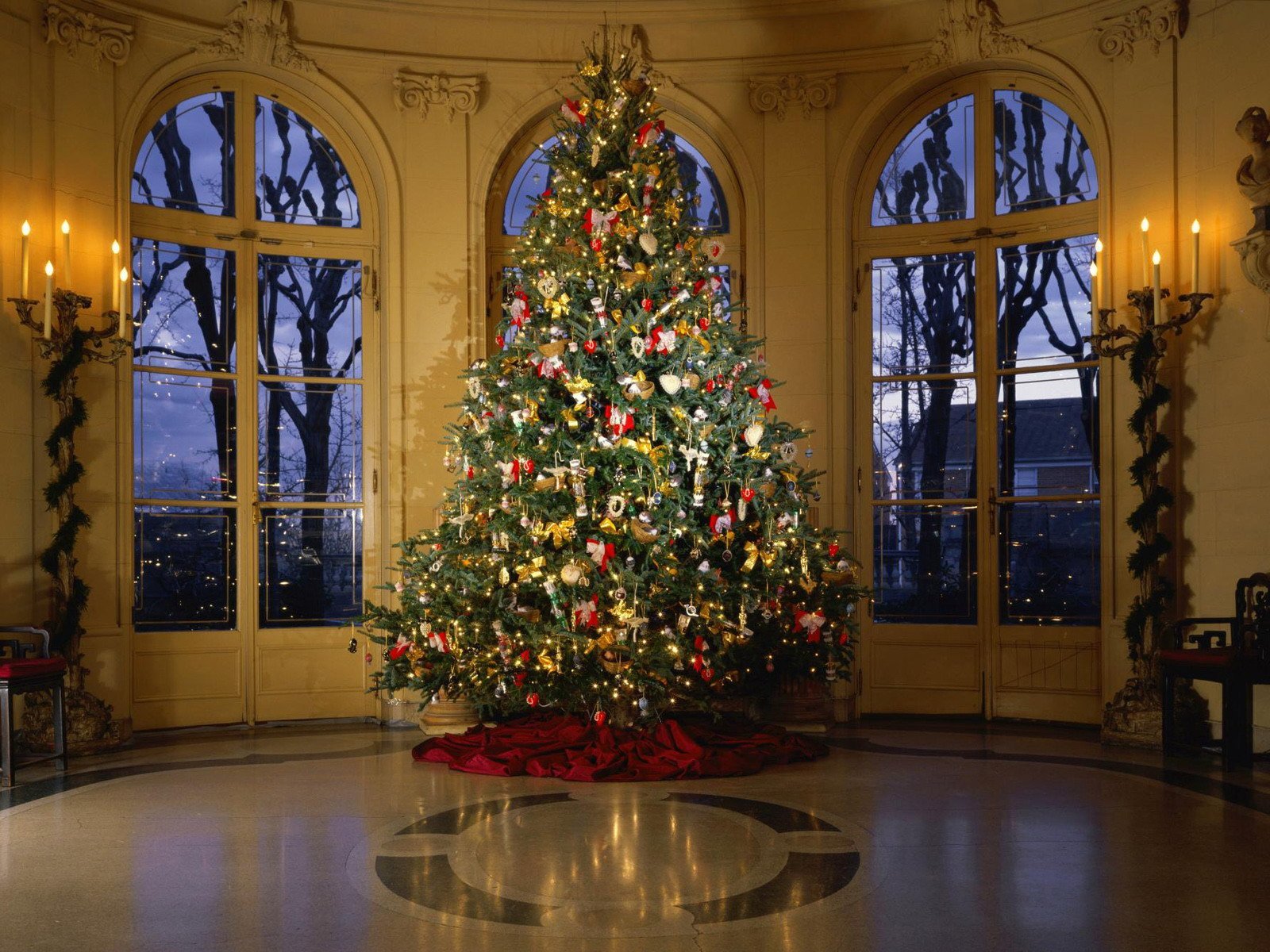 A beautifully decorated Christmas tree adorned with ornaments stands elegantly in a well-lit room, creating a festive holiday atmosphere, complemented by large windows revealing a scenic view.