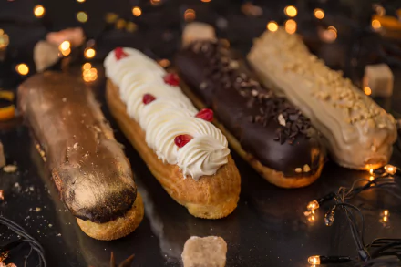 HD desktop wallpaper featuring an assortment of éclairs with varied toppings, showcasing elegant pastries and desserts on a reflective surface with warm, blurred lights.