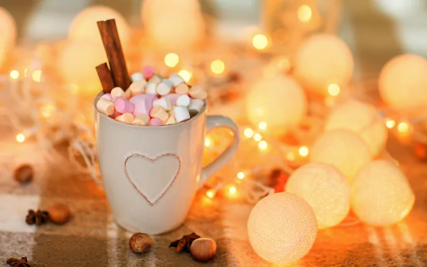 A 4K Ultra HD desktop wallpaper featuring a mug of hot chocolate topped with marshmallows and cinnamon sticks, surrounded by warm, glowing decorative lights.