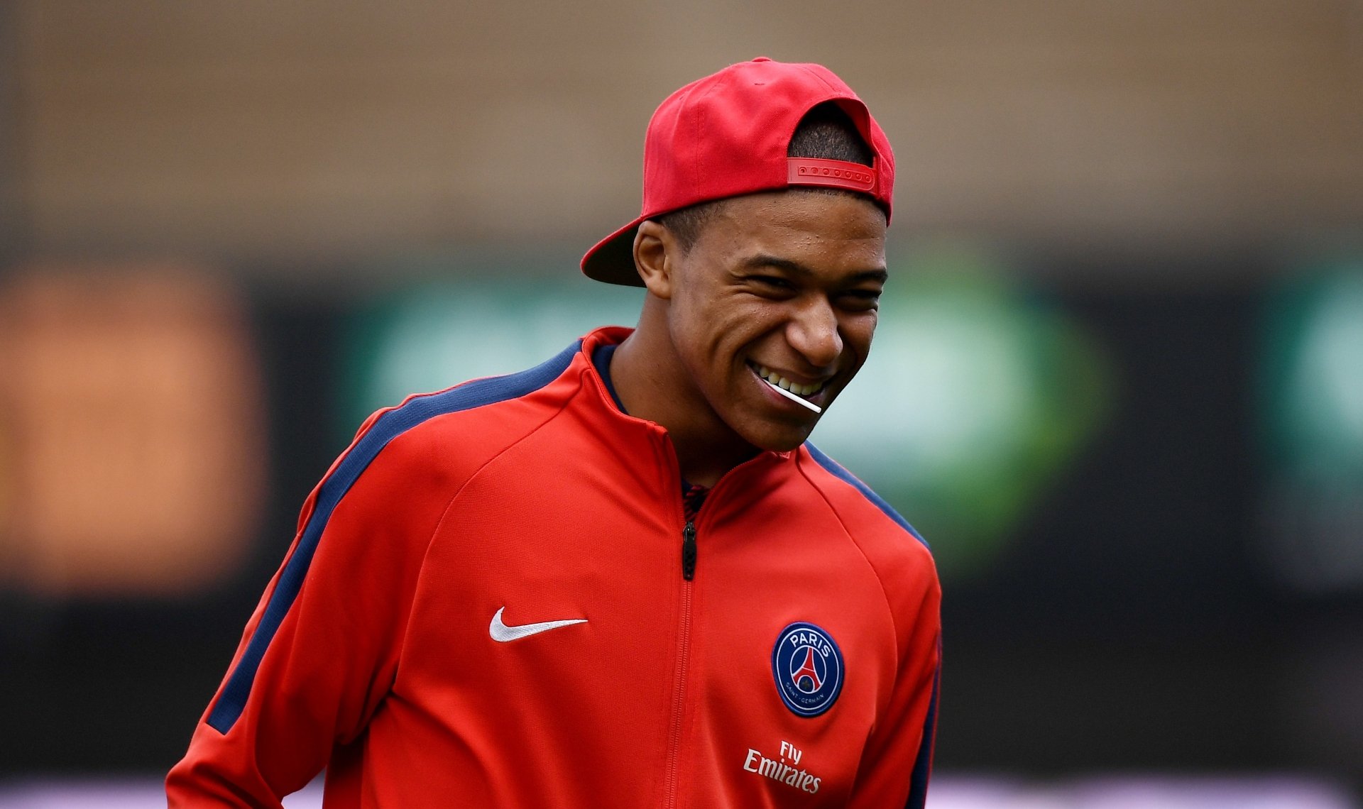 HD desktop wallpaper featuring French soccer star Kylian Mbappé smiling in a red Paris Saint-Germain jacket and cap during a sports event.