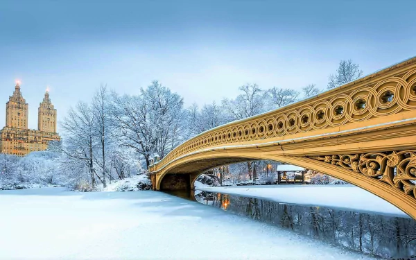 Winter view of Bow Bridge in Central Park, New York, with snow-covered trees and the city skyline in the background, captured in a high-definition desktop wallpaper.