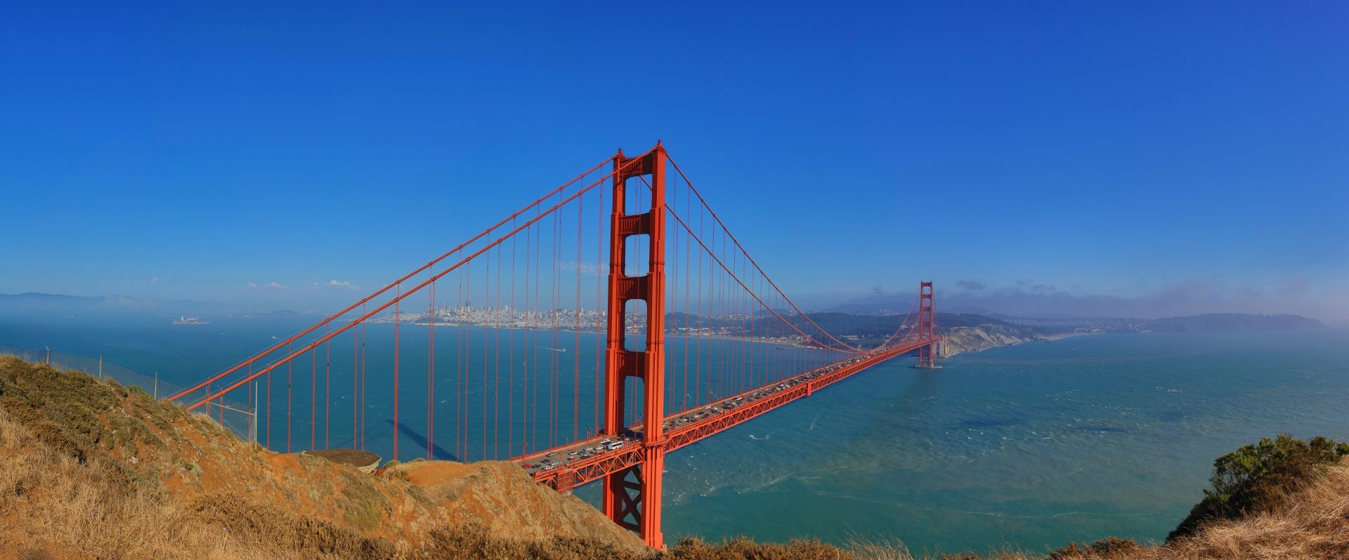 8K Ultra HD desktop wallpaper of the man-made Golden Gate Bridge spanning San Francisco Bay under a clear blue sky.