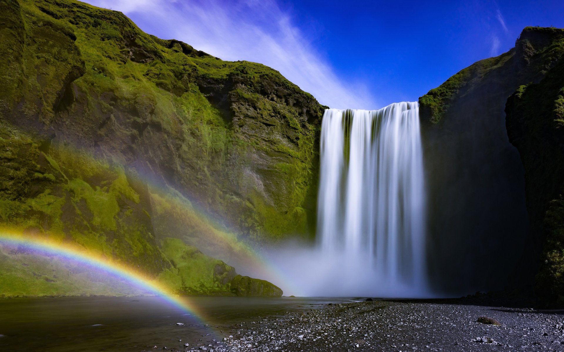 2K Quad HD PC desktop wallpaper: waterfall cascading down a mossy cliff with mist and a vibrant double rainbow over a rocky shoreline beneath a vivid blue sky.