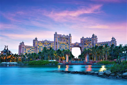 Beachfront view of the Atlantis Paradise Island hotel in Nassau, Bahamas, framed by palm trees and vibrant tropical ocean waters under a colorful sunset sky.