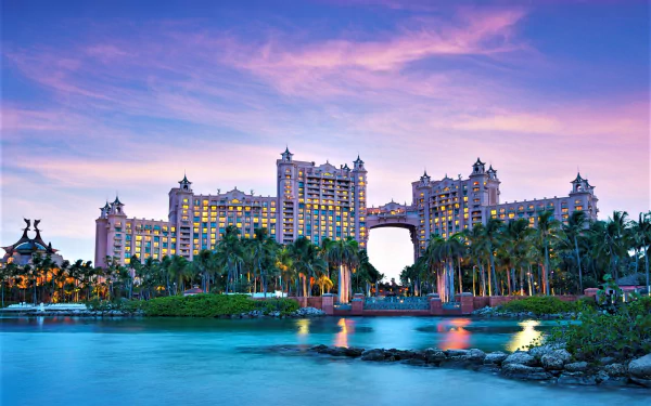 Beachfront view of the Atlantis Paradise Island hotel in Nassau, Bahamas, framed by palm trees and vibrant tropical ocean waters under a colorful sunset sky.