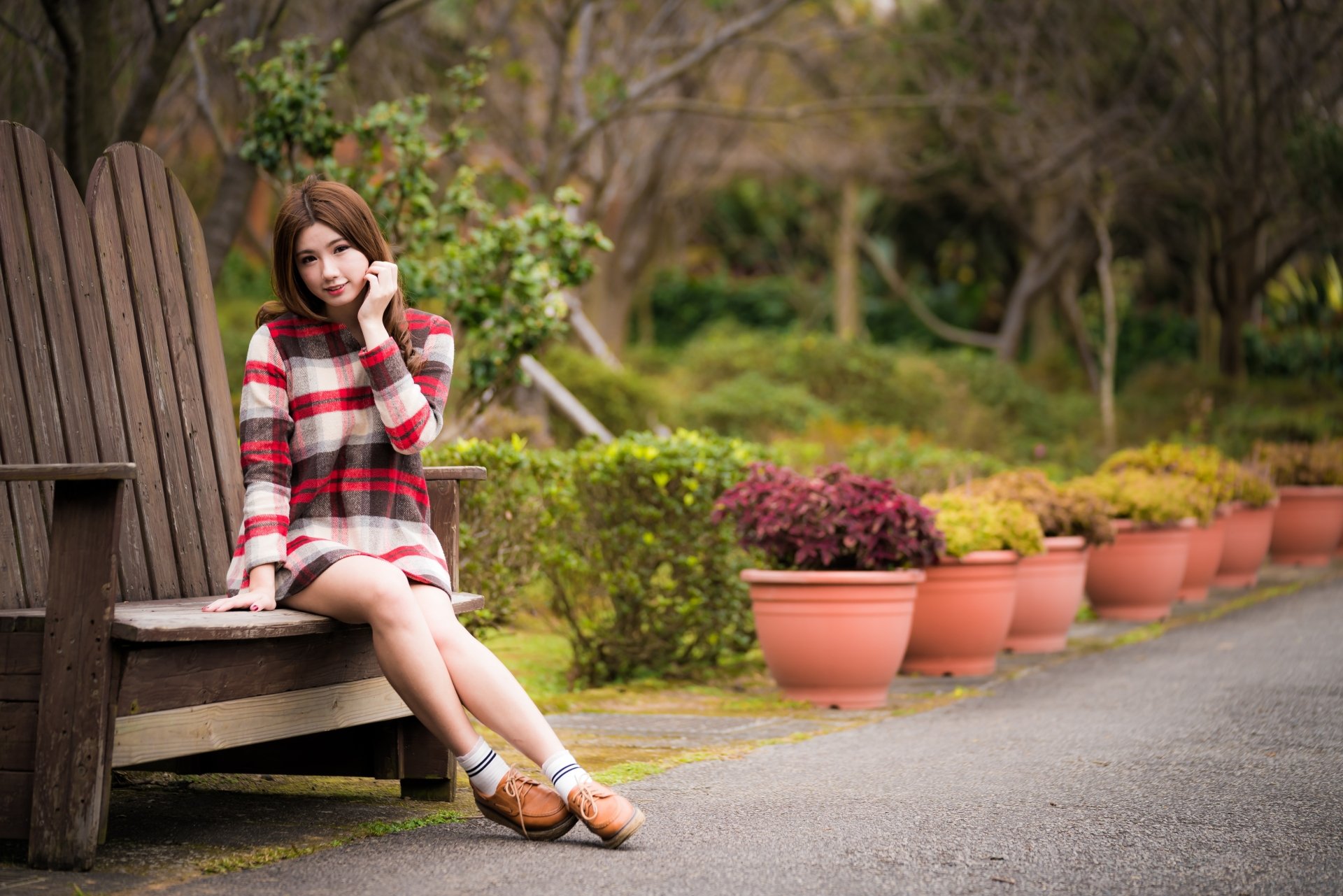 Brunette Asian woman model in a red plaid dress sits on a wooden bench surrounded by potted plants, captured with a soft depth of field in a 4K Ultra HD wallpaper.