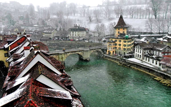 Snowfall blankets the historic houses and canal in Bern, Switzerland, showcasing a serene cityscape with a stone bridge and snowy hillside in the background.