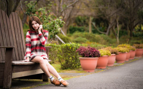 Brunette Asian woman model in a red plaid dress sits on a wooden bench surrounded by potted plants, captured with a soft depth of field in a 4K Ultra HD wallpaper.