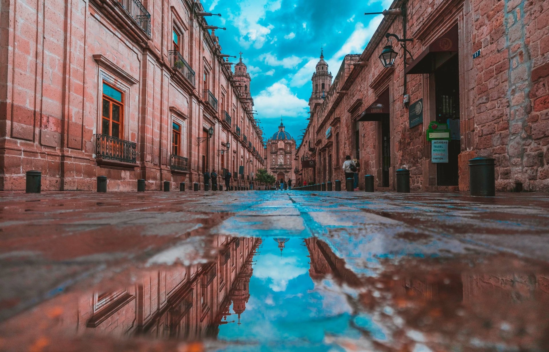 A vibrant Mexico street scene captured in HD photography, showcasing detailed reflections of historic buildings and a blue sky on water-covered cobblestones.