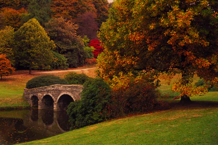 HD desktop wallpaper depicting a serene fall landscape with a stone bridge over a calm stream, surrounded by vibrant autumn trees.