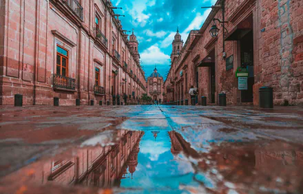 A vibrant Mexico street scene captured in HD photography, showcasing detailed reflections of historic buildings and a blue sky on water-covered cobblestones.