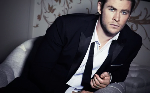 HD desktop wallpaper of Australian actor Chris Hemsworth with blue eyes, dressed in a black suit, sitting on a chair in a stylish indoor setting.