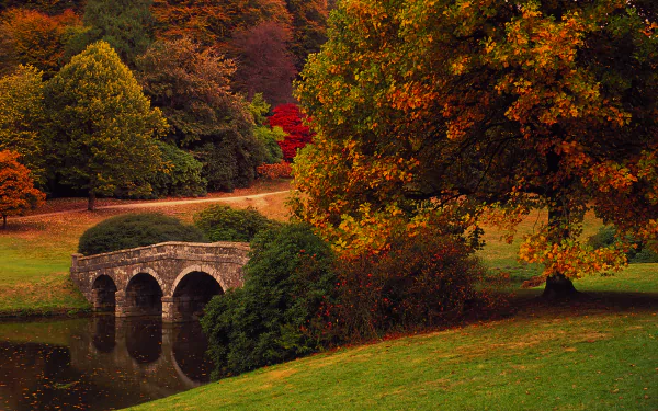 HD desktop wallpaper depicting a serene fall landscape with a stone bridge over a calm stream, surrounded by vibrant autumn trees.
