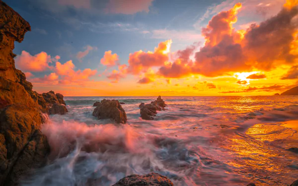8K Ultra HD desktop wallpaper of a vibrant sunset over an ocean horizon, featuring dramatic clouds and waves crashing against rocky cliffs in a stunning natural seascape.