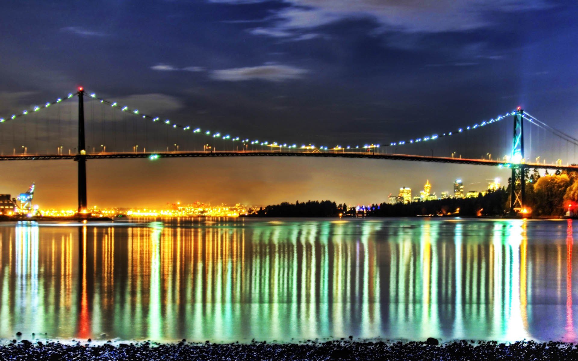Night view of the illuminated Lions Gate Bridge in Vancouver, Canada, with city lights reflecting on the calm water below.