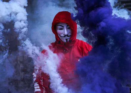 A striking HD wallpaper featuring a figure in a Guy Fawkes mask, shrouded in colorful smoke, symbolizing anonymity and technology.